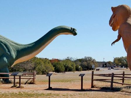 The image shows large statues of a green sauropod dinosaur and an orange theropod dinosaur in a park setting, surrounded by informational plaques and fencing.