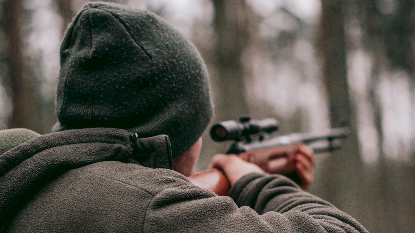 A person wearing a beanie and jacket aims a rifle in an outdoor setting with a blurred forest background.