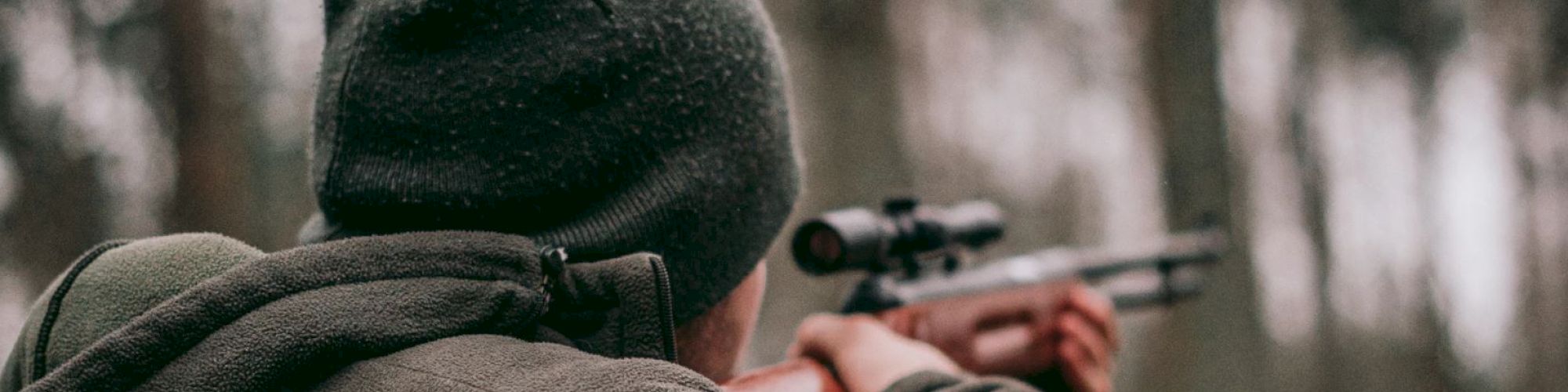 A person wearing a beanie and jacket aims a rifle in an outdoor setting with a blurred forest background.