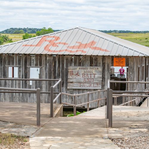 A rustic wooden boathouse with a metal roof sits by a lake, surrounded by greenery, featuring a sign and a ramp.