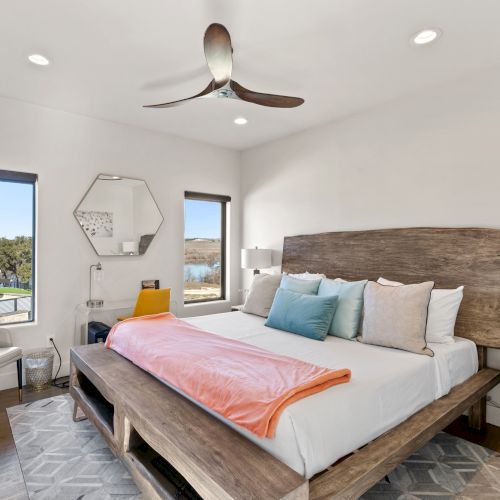 A modern bedroom with a large bed, colorful pillows, an orange throw, two windows, a chair, a mirror, and a ceiling fan.