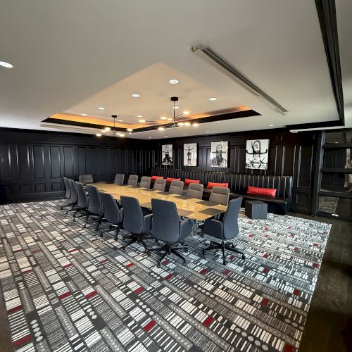 The image shows a modern conference room with a long table, several chairs, patterned carpet, dark walls, and framed art.