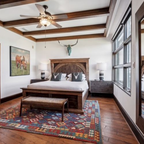 A cozy bedroom features a wooden bed, colorful rug, ceiling fan, window with light, and artwork of cows and horns above the bed.