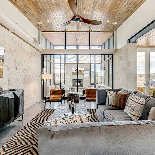 A modern living room with a wooden ceiling, grey couches, a large TV, and floor-to-ceiling windows offering a scenic view outside.