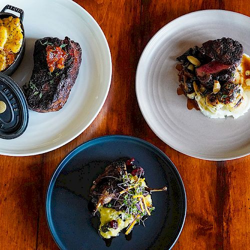 The image shows three plates of gourmet food, each featuring different meat dishes, garnishes, and sauces, arranged on a wooden table.