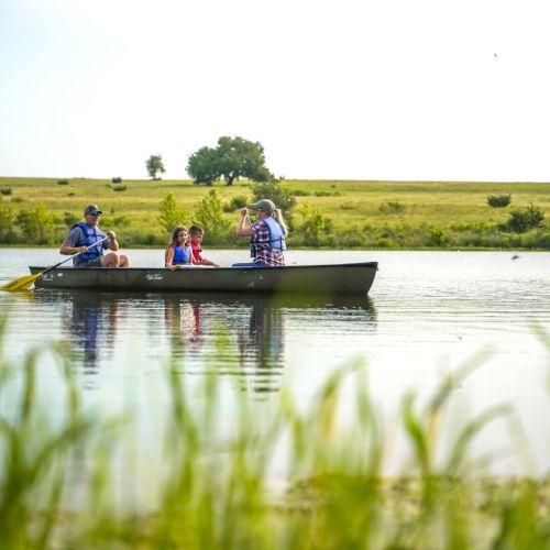 A family is canoeing on a calm lake surrounded by greenery, under a clear sky.