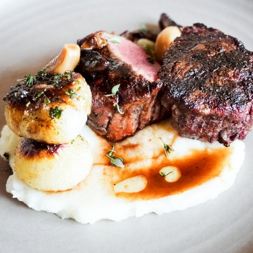 The image shows a gourmet dish with seared meat, possibly lamb or beef, on a bed of creamy mashed potatoes, garnished with herbs and sauce.