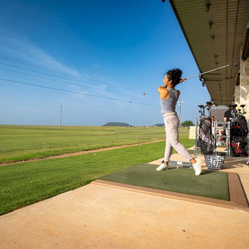 A person is swinging a golf club at a driving range, with golf bags and a vast green field in the background.