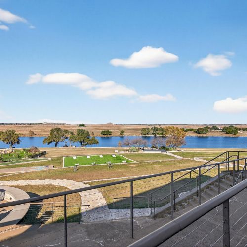 A sunny coastal scene with a rocky shoreline, clear blue sky, gentle sea, and a railing along a boardwalk.