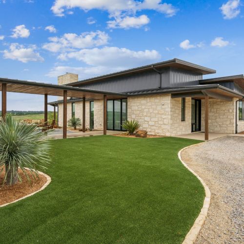 A modern house with stone and metal exterior, covered patios, green lawn, and gravel driveway under a blue sky with clouds.