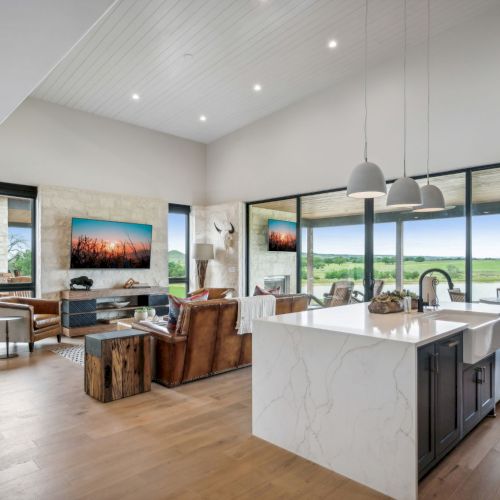 A modern living area with a kitchen island, pendant lights, large windows, and cozy seating overlooking a scenic view, completes the decor.