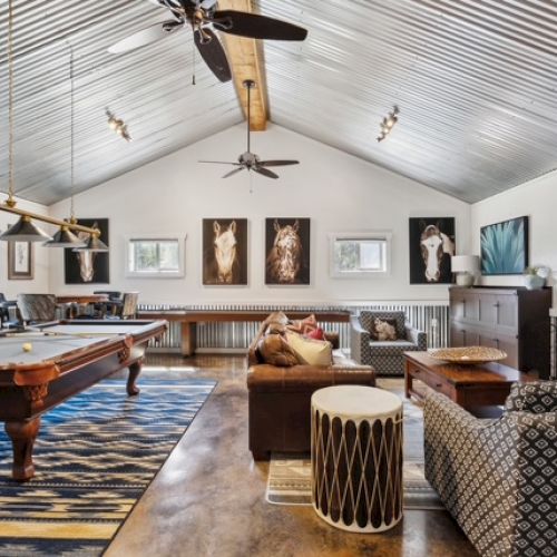 A cozy room with a pool table, sofas, artwork on the walls, a patterned rug, and a fireplace under a high, corrugated metal ceiling.