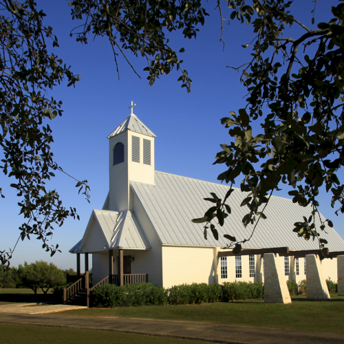 A quaint white church with a pitched roof and bell tower is nestled among trees under a clear blue sky.