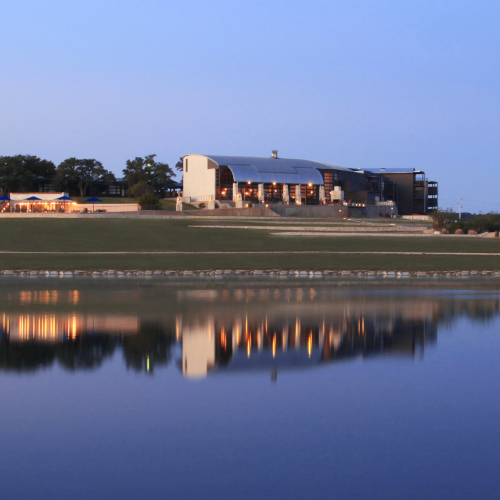 A modern building sits by a calm lake, reflecting on the water, with a clear blue sky and surrounding greenery completing the scene.
