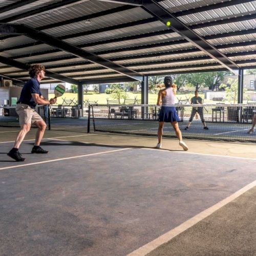 People playing pickleball indoors on a court with a metal roof. Two teams of two players are actively engaged in a match.