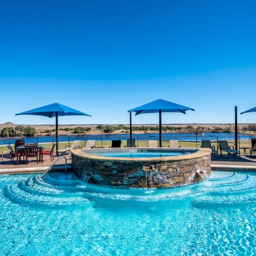 A swimming pool with an elevated spa and cascading water, surrounded by lounge chairs and blue umbrellas, overlooks a scenic lake.