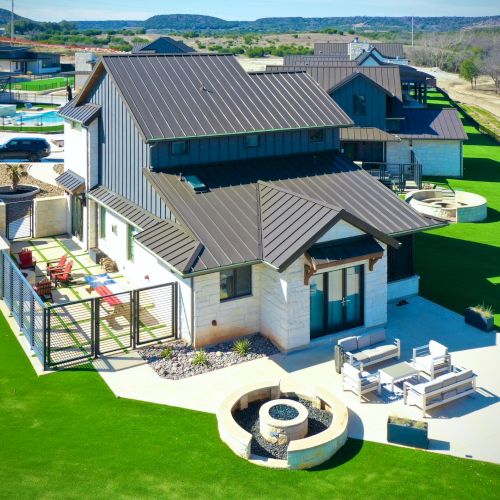 A modern house with a dark roof, surrounded by lush green lawns. It features outdoor seating areas, a fire pit, and a pool.