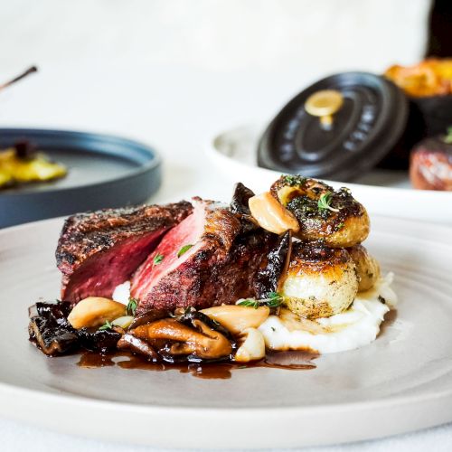 The image shows a plated dish with sliced steak, roasted vegetables, and garnish, accompanied by other dishes in the background.