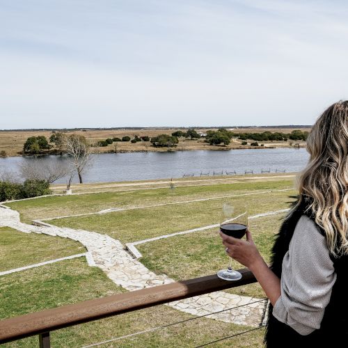 A person with wavy hair holds a drink while looking at a scenic view of a river and a path through grassy fields from a balcony.