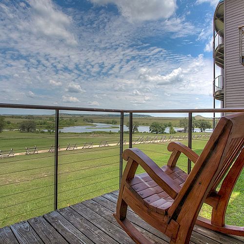 The image shows a wooden rocking chair on a balcony overlooking a scenic landscape with green fields, a body of water, and a partly cloudy sky.