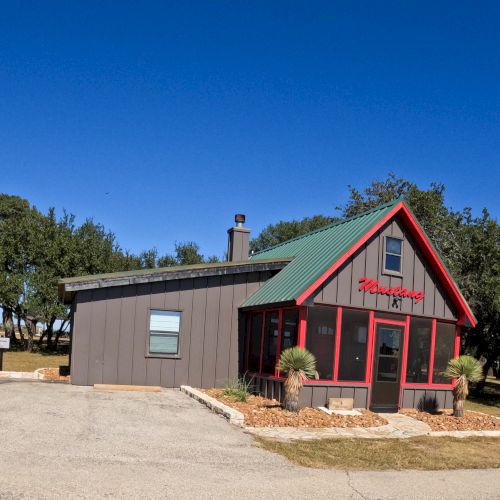The image shows a small, cabin-like building with red trim and a green roof on a sunny day, surrounded by trees and a gravel driveway.