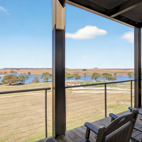 A sunny balcony view over the beach and sea with a wooden railing and cushioned seating, framed by a shaded terrace and clear blue sky.