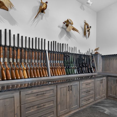 A gun rack with dozens of rifles arranged in a U-shaped display, matching wooden cabinets, and mounted animal heads on the white wall.