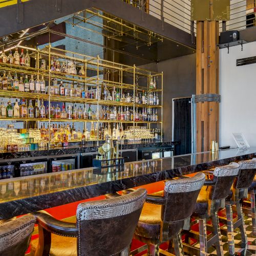 A stylish bar with a long marble counter, high chairs, and shelves stocked with bottles in the background, under modern decor.