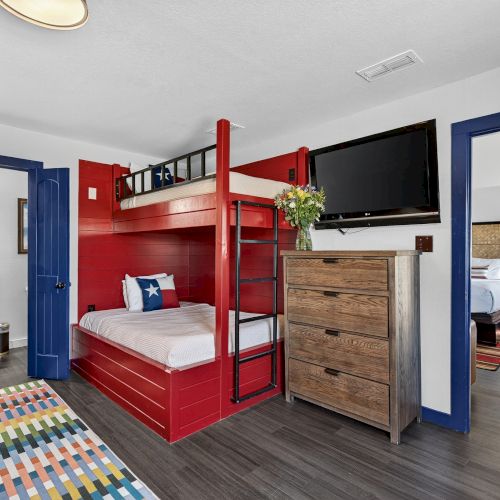 A bedroom with a red bunk bed, colorful rug, dresser, TV, and en suite bathroom featuring red cabinetry and blue trim accents.