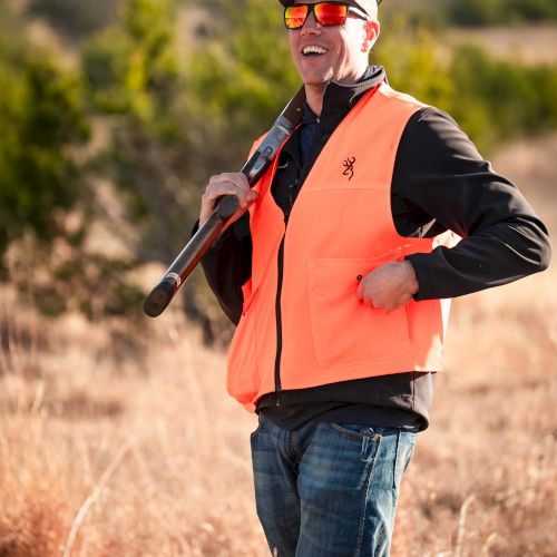 A man in an orange safety vest with sunglasses and a cap holds a shotgun over his shoulder in a dry, grassy outdoor area.