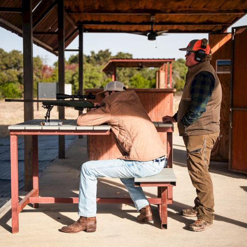A man in a chair at a shooting bench aims a rifle while another man watches, outdoors at a shooting range with wooden booths and signs.