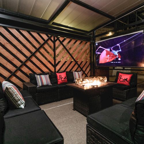 A modern, dimly lit indoor event space with a wooden lattice wall, stage, large screens, and a few attendees seated in front of a speaker setup, audience facing stage.