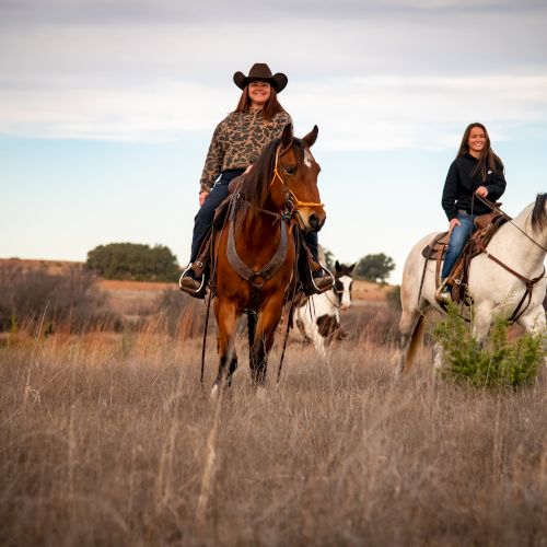 Three people horseback riding through a wide grassy plain, one in a leopard print jacket, the others in dark jackets, at sunset.