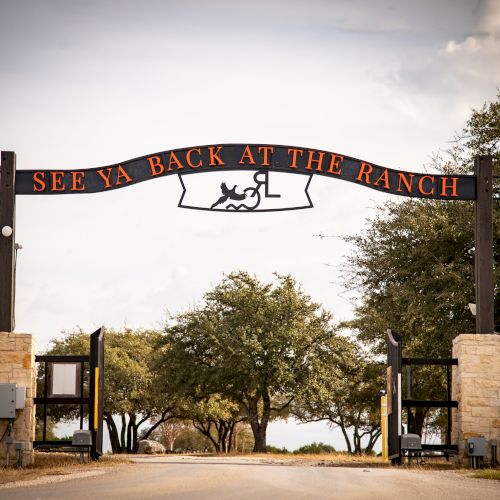 A rustic gated entrance to a ranch with a curved sign reading &ldquo;SEE YA BACK AT THE RANCH,&rdquo; flanked by stone pillars and trees in the background.