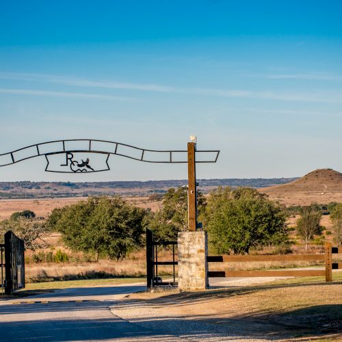 The image shows a ranch entrance with a metal gate and a sign, set against a backdrop of open fields and hills under a clear blue sky.