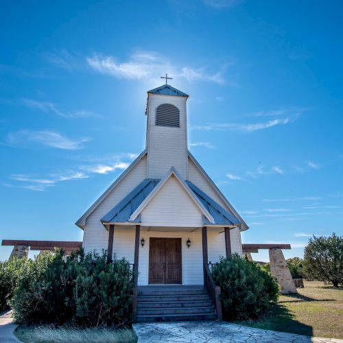 A small, white chapel with a steeple and cross, surrounded by bushes, stands under a clear blue sky in a grassy landscape.