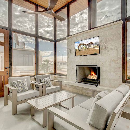 A cozy sunroom features modern furniture, a fireplace, and a wall-mounted TV, with large windows offering an expansive outdoor view.