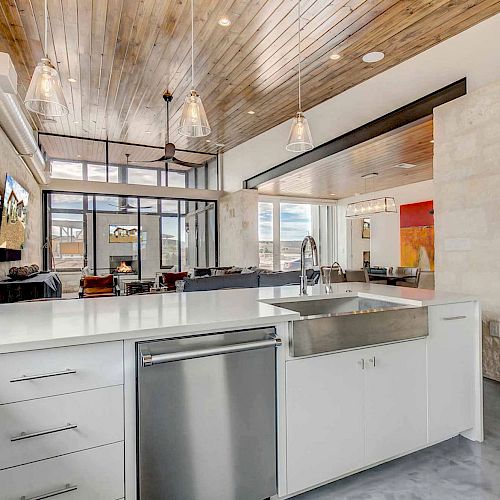 A modern kitchen with white cabinets, stainless steel appliances, and a large island with a sink; open layout with wood ceiling and pendant lights.