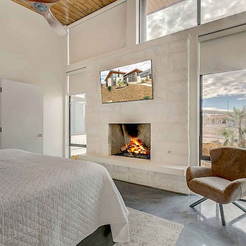 A modern bedroom features a cozy fireplace, wall-mounted TV, large windows, white bedding, a brown chair, and a view of an outdoor scene.