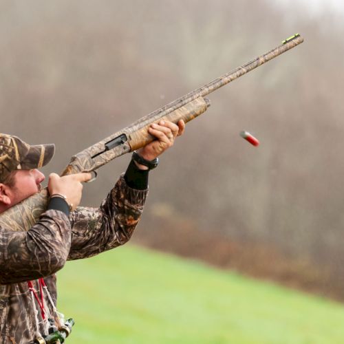 A person in camouflage clothing is firing a shotgun outdoors, with a shell casing visible in the air.