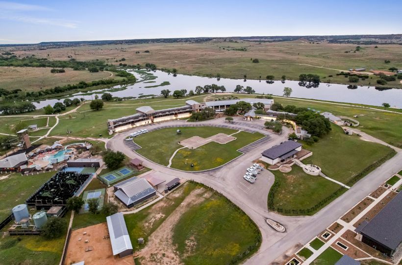 Aerial view of a countryside lodge/resort with pools, sports fields, parking cars, and a winding river running nearby.