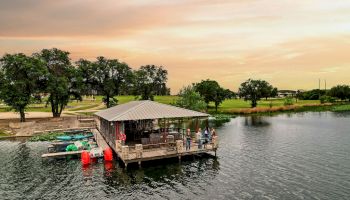 A lakeside dock with a covered boat shelter, kayaks, and people gathered by the water at sunset.