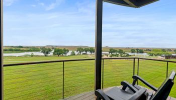 A riverside balcony with patio chairs overlooks a green field, trees, and a calm river under a blue sky.