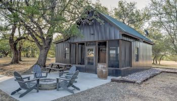 The image shows a small metal cabin with a green roof, surrounded by trees, featuring a fire pit with chairs and a barrel outside.