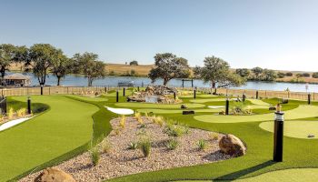 A beautifully landscaped mini-golf course by a lakeside, with lush greens, rocks, and trees under a clear blue sky.