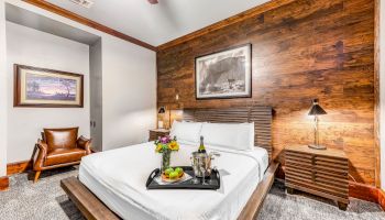 A modern bedroom features a wooden accent wall, a bed with white linens, nightstands, framed artwork, a leather chair, and a tray with a bottle and fruits.