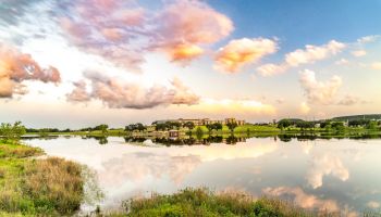 A tranquil lake scene at sunset with reflections of the sky and clouds, surrounded by grass and trees, with buildings in the background.