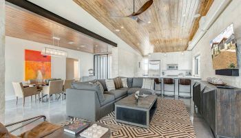 Modern living room with wooden ceiling, gray sectional sofa, geometric rug, dining area with colorful artwork, and open kitchen layout.