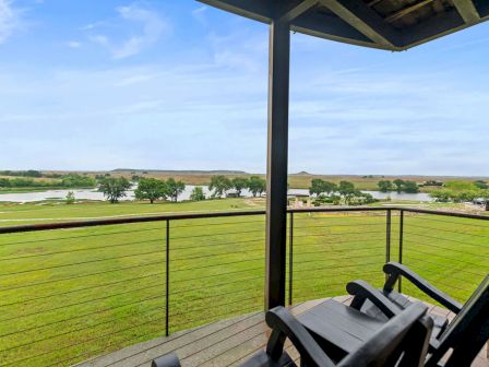 A lakeside porch with chairs overlooking a green pasture, a slow river, and distant hills under a blue sky.