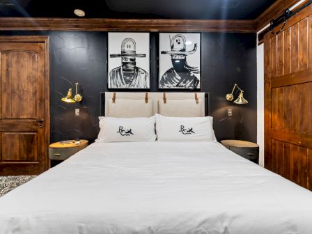 A modern bedroom with a white double bed, wooden doors, brass lamps, and two black-and-white gun-themed wall artworks above the headboard.
